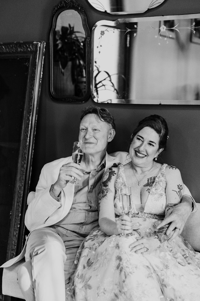 Black and white photo of smiling couple in formal attire sitting together, holding champagne glasses, in front of vintage mirrors at Firehouse Chicago