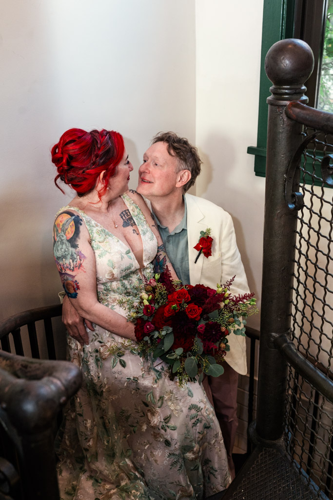 Bride and groom sit close together on a staircase at Firehouse Chicago, smiling and holding a bouquet of red flowers