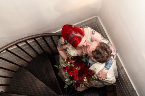 A couple embraces and kisses on a spiral staircase at Firehouse Chicago, holding a bouquet of red flowers.