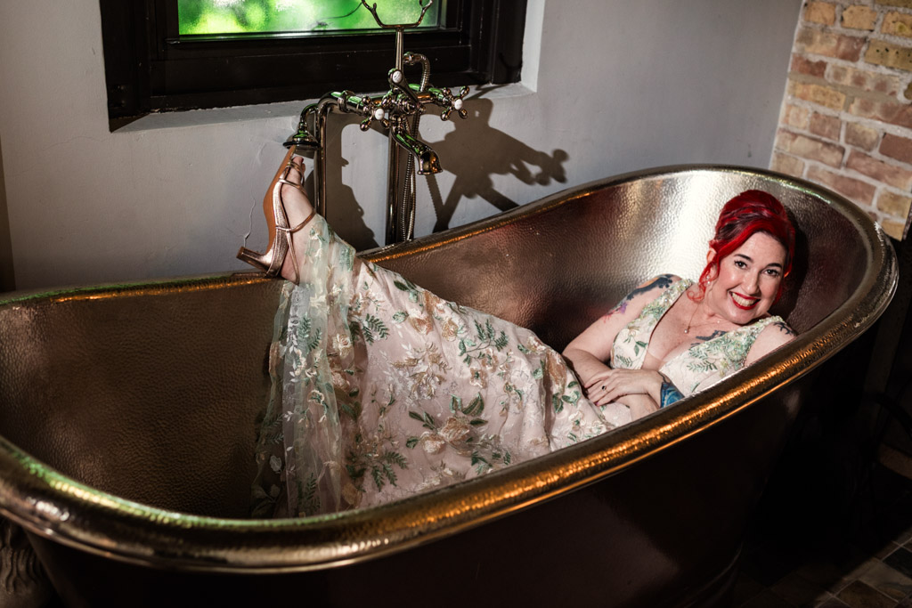 Bride with red hair in a floral dress lounges in a vintage bronze bathtub at Firehouse Chicago