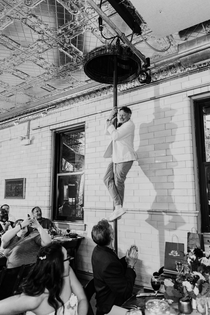 Black and white photo of groom sliding down a fire pole indoors at Firehouse Chicago as seated guests watch and applaud