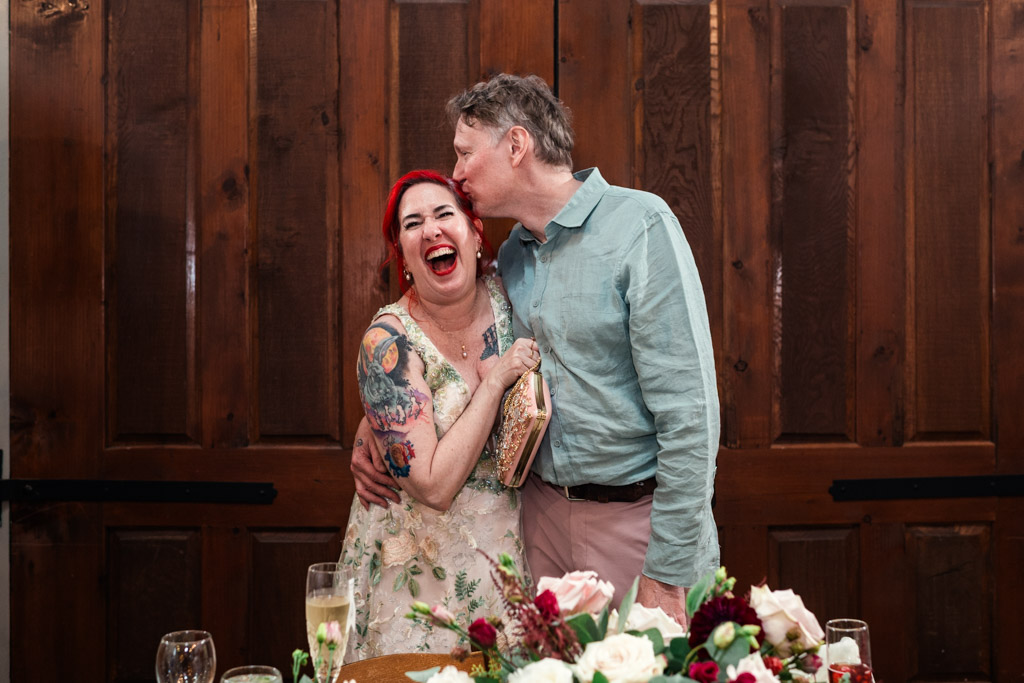 A man kisses a laughing woman with red hair and tattoos, standing in front of the wooden doors at Firehouse Chicago during a celebration.