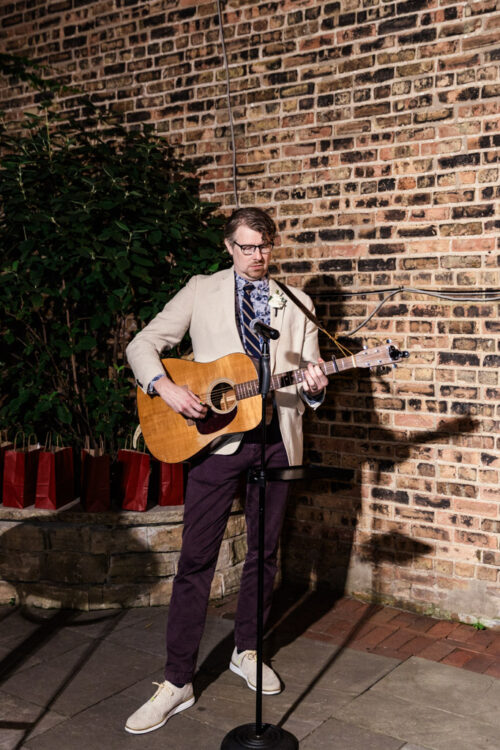 Man in a beige blazer playing an acoustic guitar and singing into a microphone outdoors at night at Firehouse Chicago.