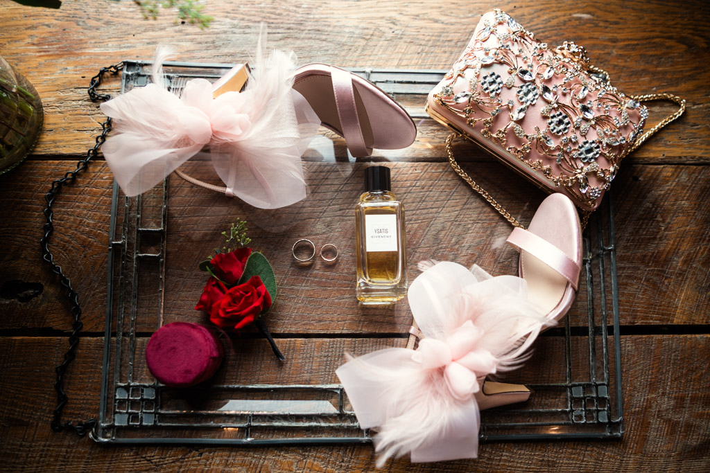 Bride's heels with bows, a floral clutch, perfume, rings, and a red boutonniere are displayed on a glass tray for wedding celebration at Firehouse Chicago
