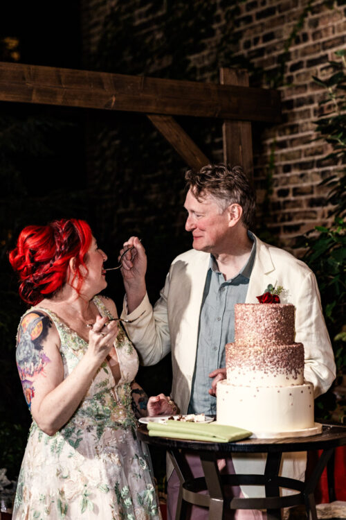 A couple smiles and feeds each other cake at their wedding celebration at Firehouse Chicago, standing beside a beautifully decorated cake.