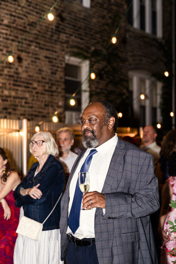 A man in a suit holding a champagne glass stands outside among guests under string lights at night during wedding reception at Firehouse Chicago