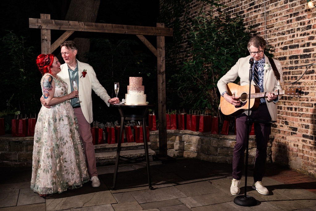 Newlywed couple stands by their wedding cake, smiling, as a man plays guitar and sings outdoors at night at Firehouse Chicago