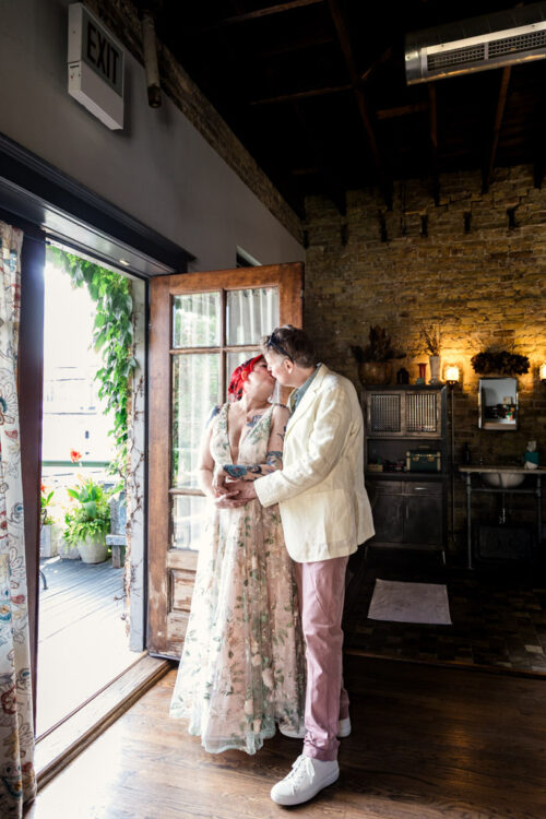 A couple dressed in wedding attire shares a kiss by an open door in a rustic, sunlit room at Firehouse Chicago.