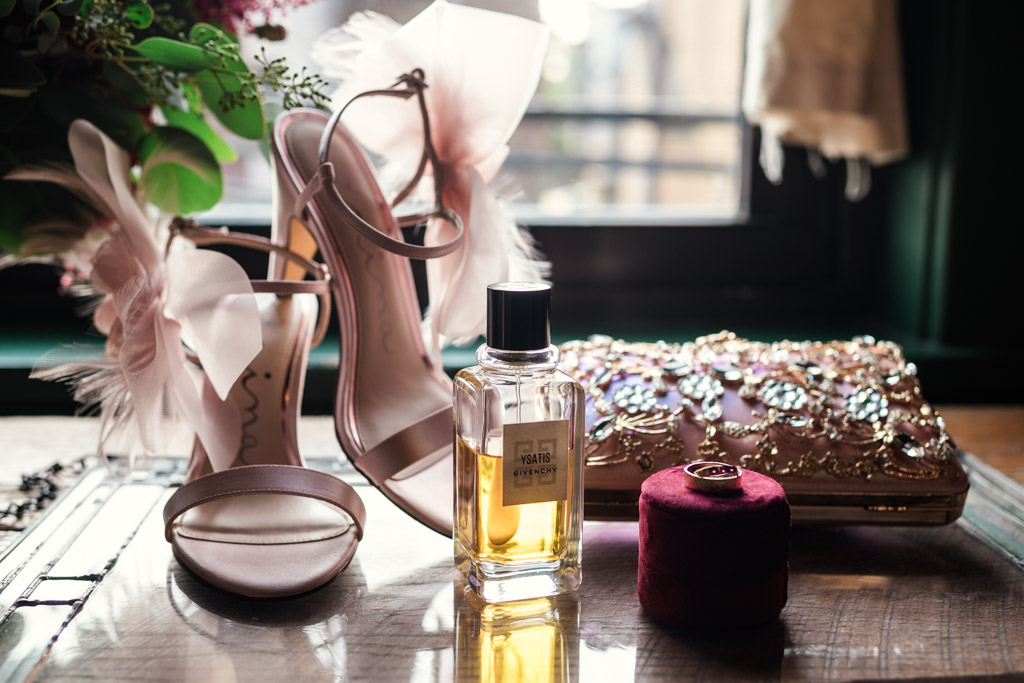 Bride's shoes, perfume, jeweled clutch, and a ring box rest on a glass table by a window for Firehouse Chicago wedding celebration