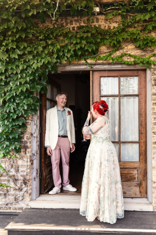 A smiling couple stands at a doorway covered in green ivy at Firehouse Chicago; the woman wears a floral dress, the man a white jacket.