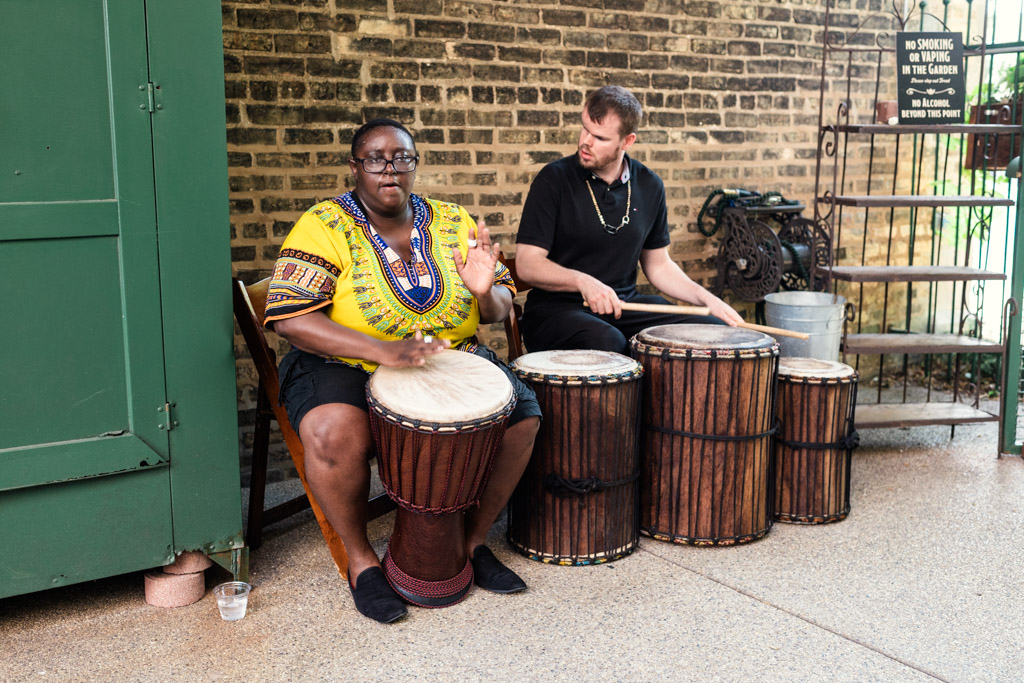 Two people sit and play hand drums beside a brick wall at Firehouse Chicago, one wearing a yellow patterned shirt