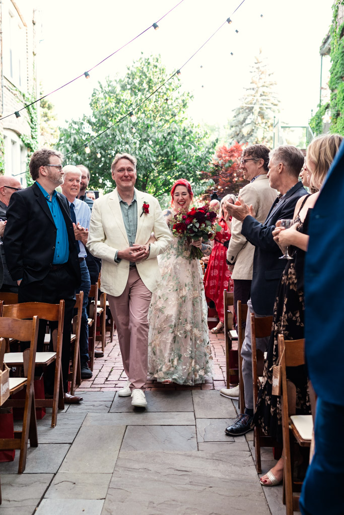 Bride with a bouquet and groom walk down the outdoor aisle at Firehouse Chicago, with guests applauding
