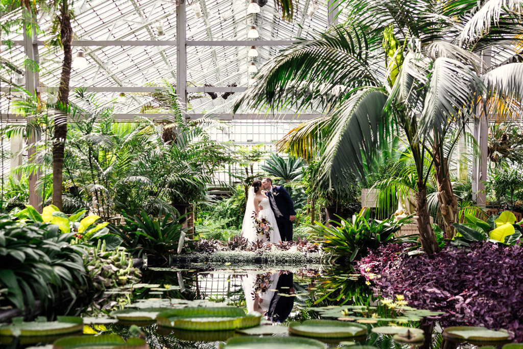 Bride and groom kiss in a greenhouse at Garfield Park Conservatory, surrounded by tropical plants and a reflective pond