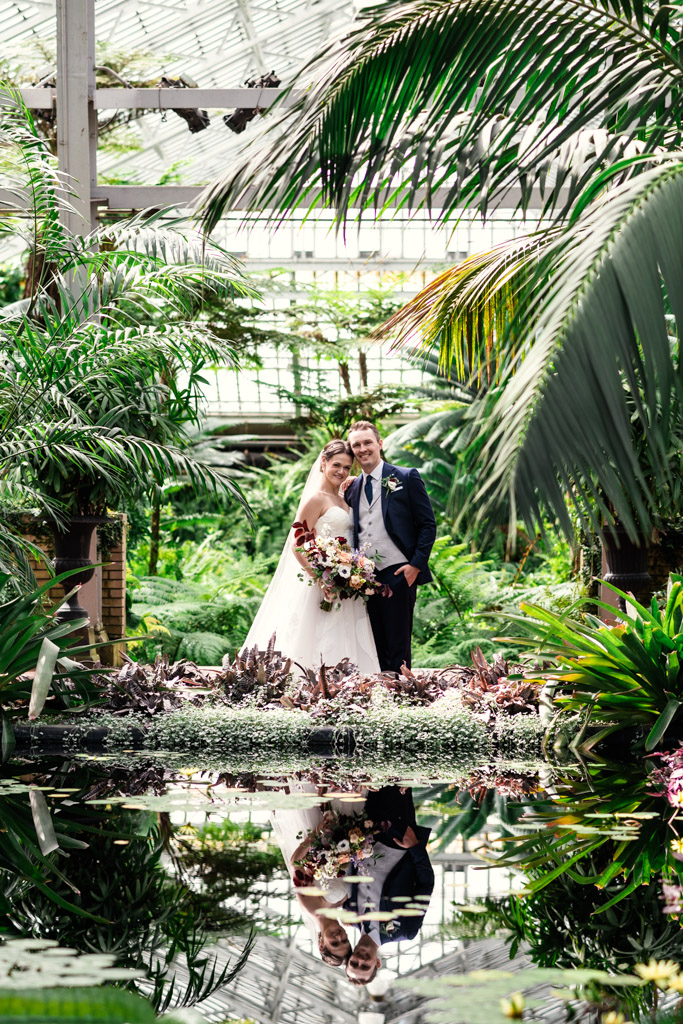 Bride and groom stand smiling together surrounded by lush green foliage at Garfield Park Conservatory, their reflections visible in a still pond