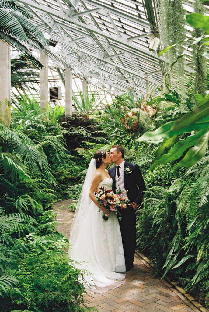 Bride and groom kiss on a brick path in a greenhouse at Garfield Park Conservatory, surrounded by ferns and greenery