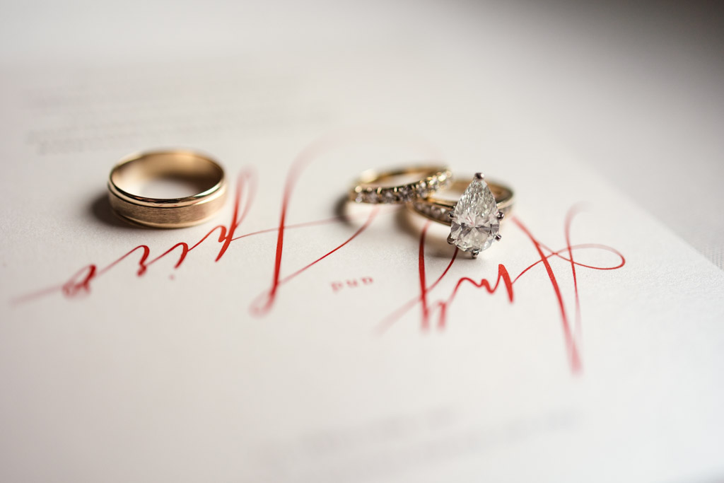 Detail photo of three gold rings, including a diamond engagement ring, rest on a save-the-date card with elegant red calligraphy for Lincolnshire Marriott Resort wedding celebration