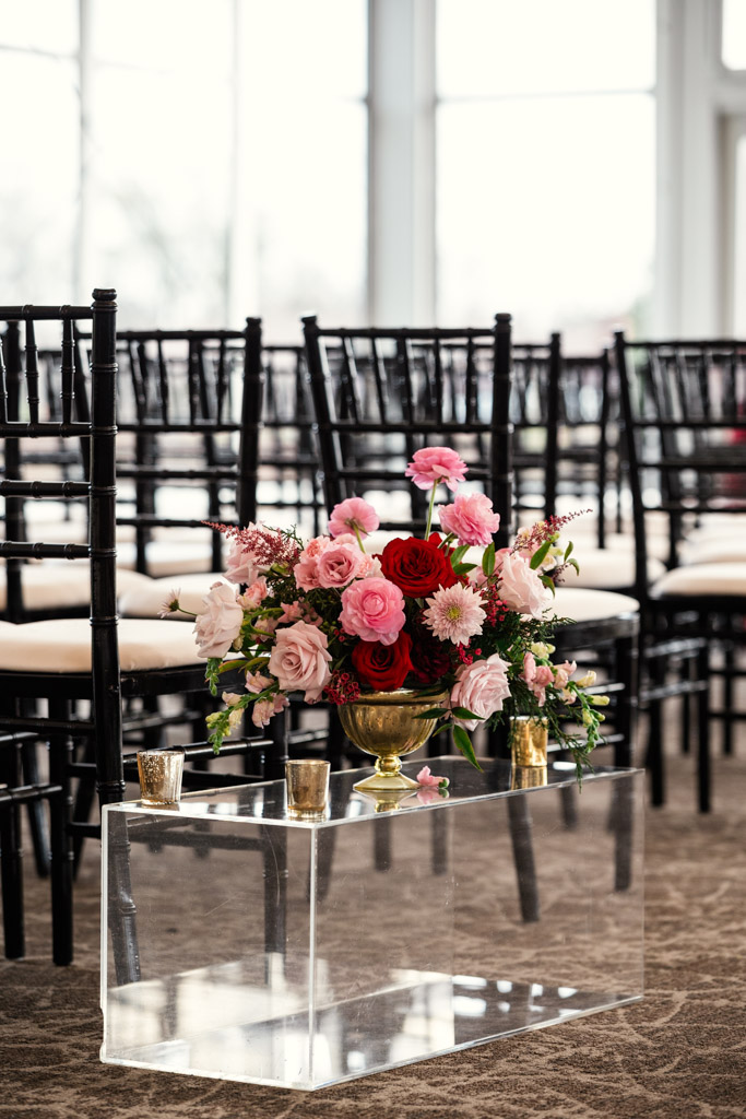 A gold vase with pink and red flowers sits on a clear stand beside rows of black chairs for wedding ceremony at Lincolnshire Marriott Resort
