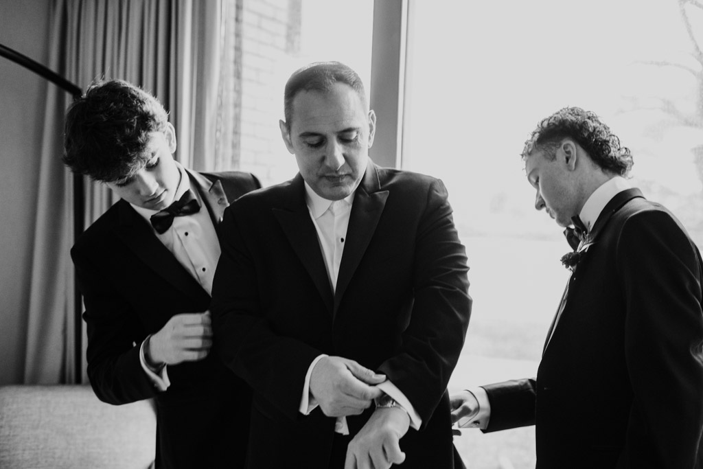 Groom and his sons in tuxedos adjust their sleeves and jackets near a window at Lincolnshire Marriott Resort