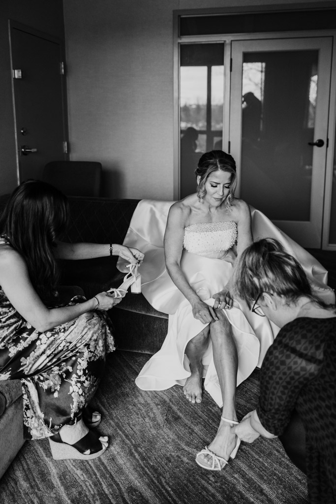 Black and white photo of bride sitting on a couch at Lincolnshire Marriott Resort while two women help her put on her shoes before the wedding