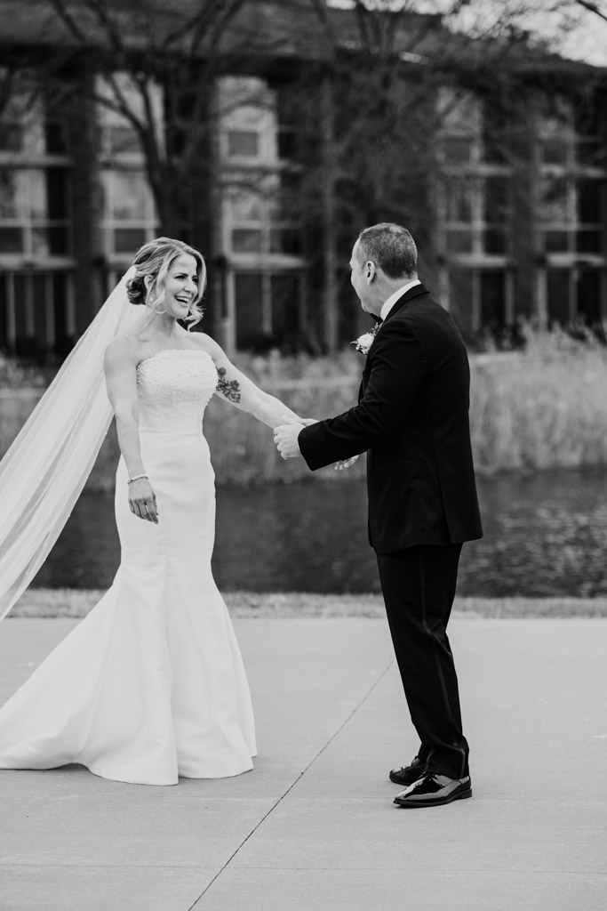 Black and white photo of bride and groom smile and hold hands outdoors by the water during their first look at Lincolnshire Marriott Resort