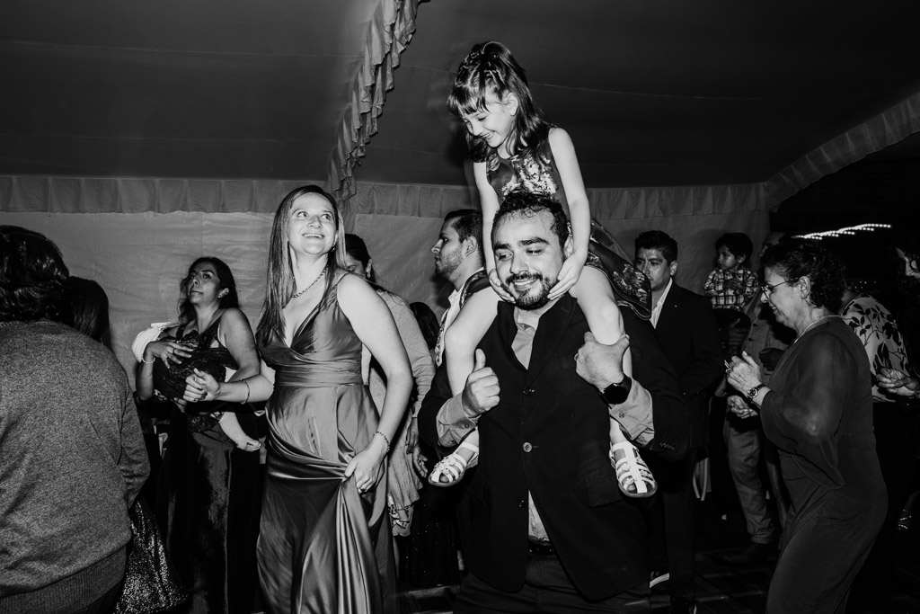 A man carries a smiling girl on his shoulders while people dance and celebrate at a lively Mexico City wedding.