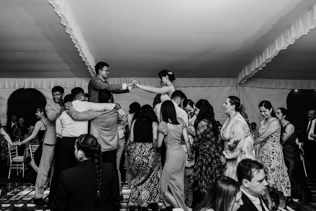 Black and white photo of bride and groom lifted on chairs by guests, during their Mexico City wedding reception