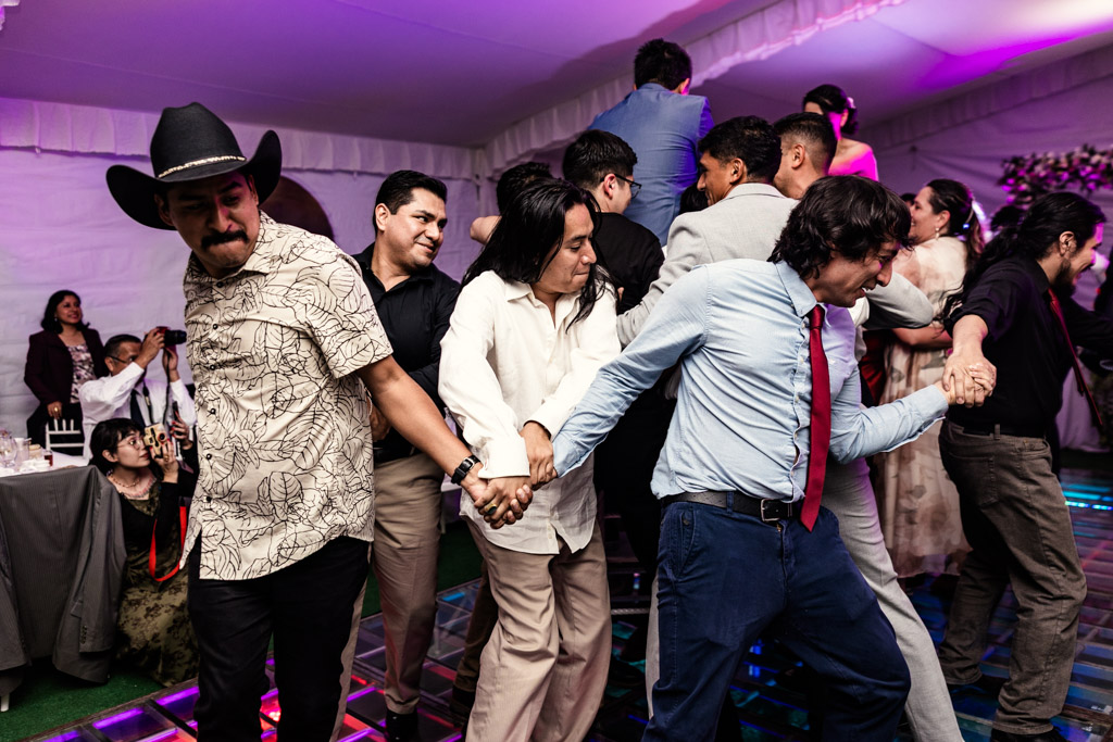 A group of people form a dancing chain at a lively indoor Mexico City wedding, holding hands and smiling.