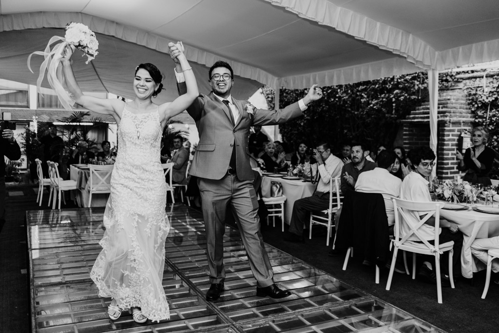 Black and white photo of bride and groom entering their Mexico City wedding reception, holding hands and smiling, as guests seated at tables watch