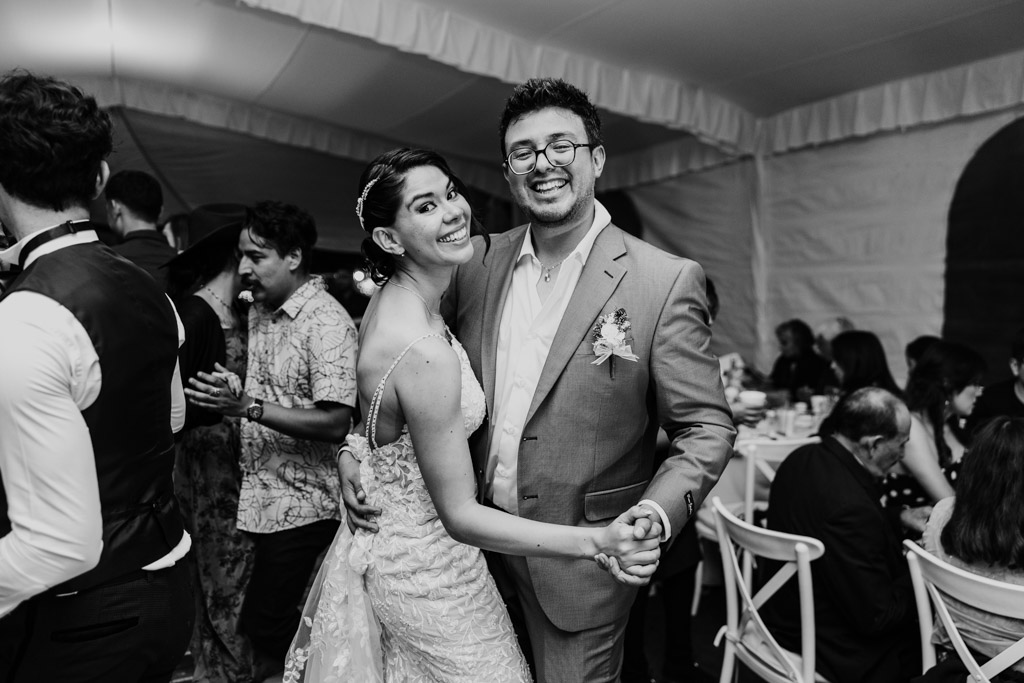 Black and white photo of newlyweds smiling while dancing together at their Mexico City wedding reception, surrounded by joyful guests