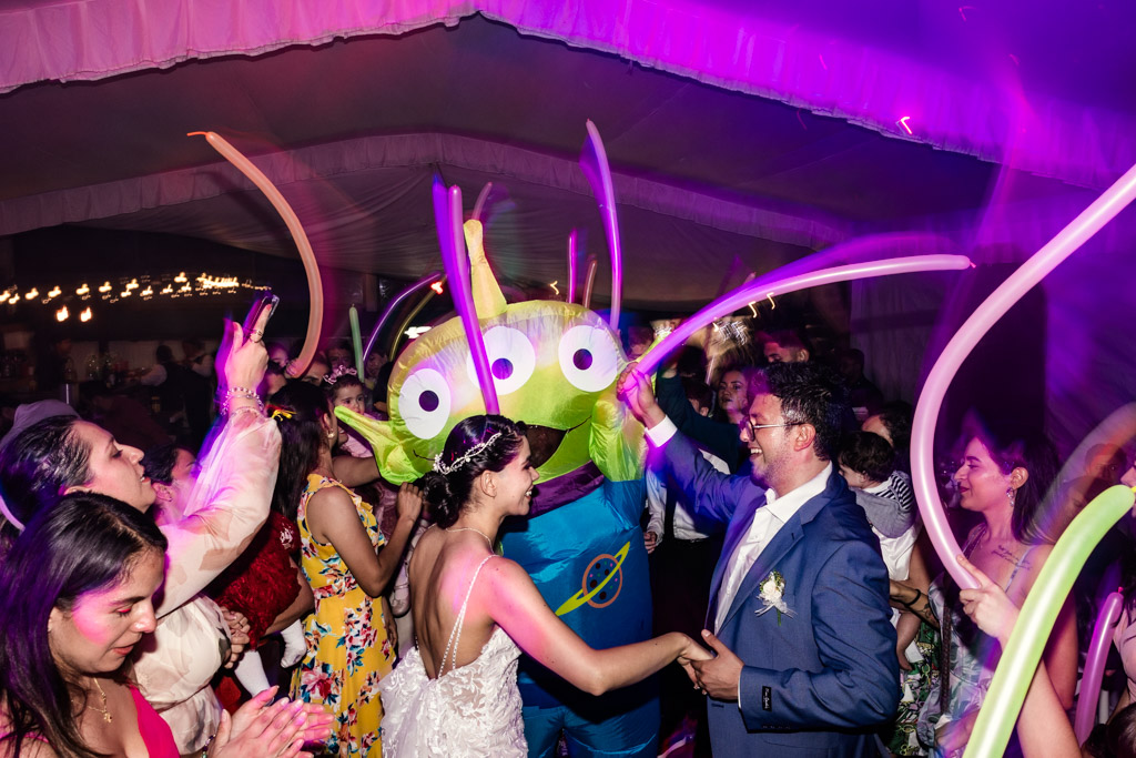 A bride and groom dance at their Mexico City wedding reception with guests and a person in a green alien costume, some people holding colorful balloons