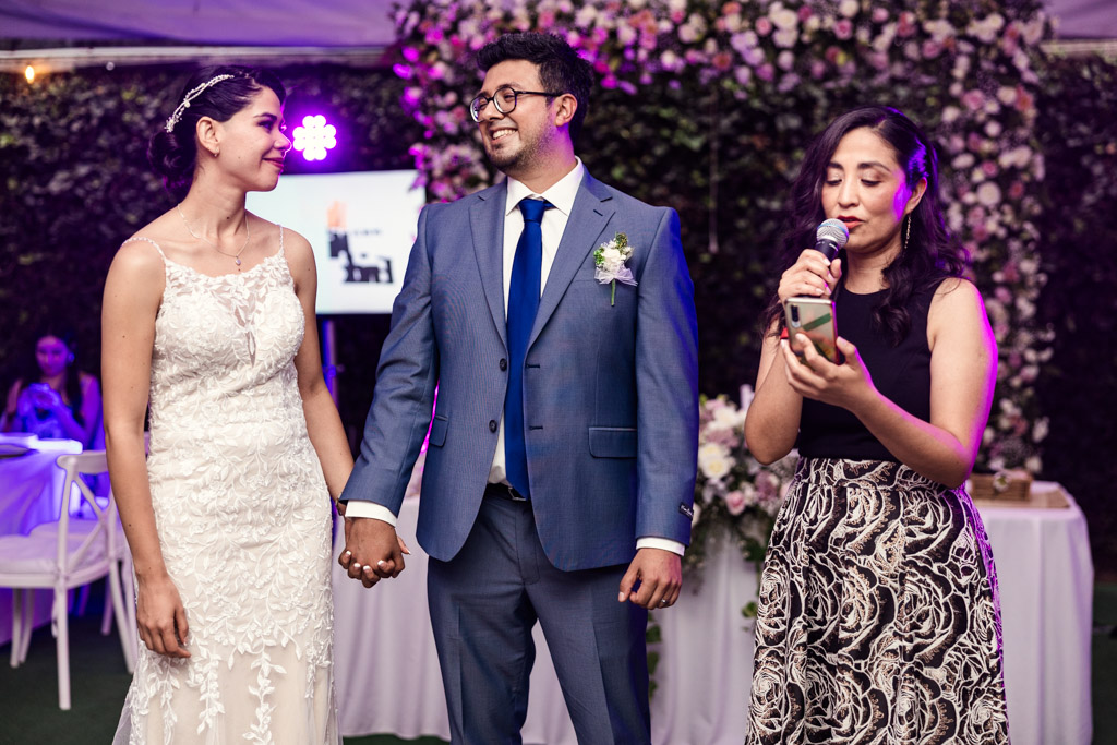 Happy bride and groom hold hands while a woman gives a speech at their Mexico City wedding reception