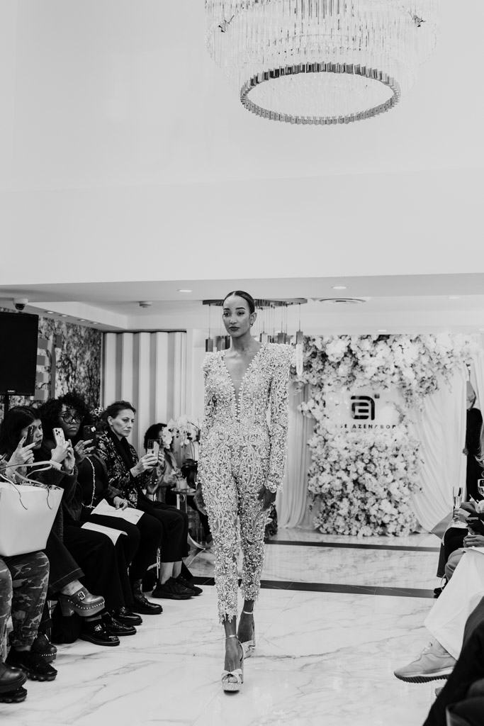 A model walks down a marble runway in a sparkling jumpsuit at New York Bridal Fashion Week, with people seated on both sides.