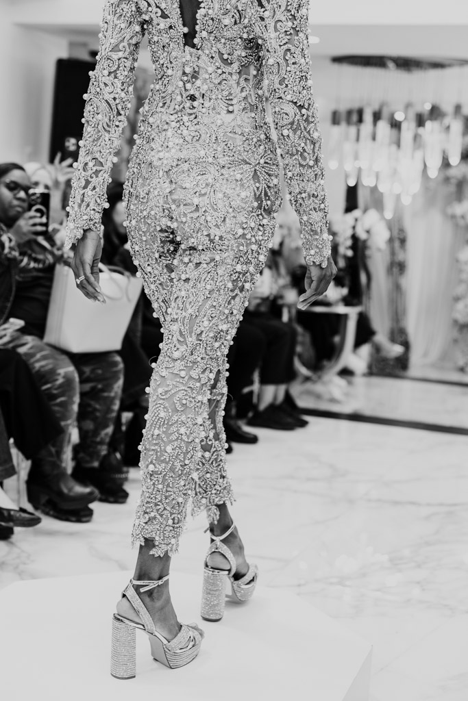 A model in an ornate, beaded jumpsuit and high heels walks the runway at New York Bridal Fashion Week, captivating a seated audience.
