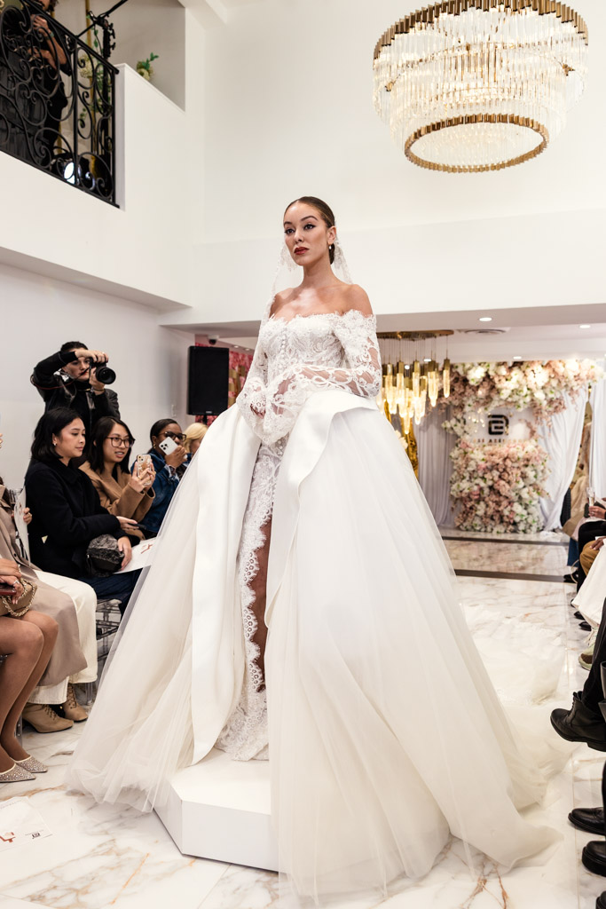 A bride in an elegant white gown stands in a decorated room surrounded by seated guests and photographers, capturing the essence of New York Bridal Fashion Week.