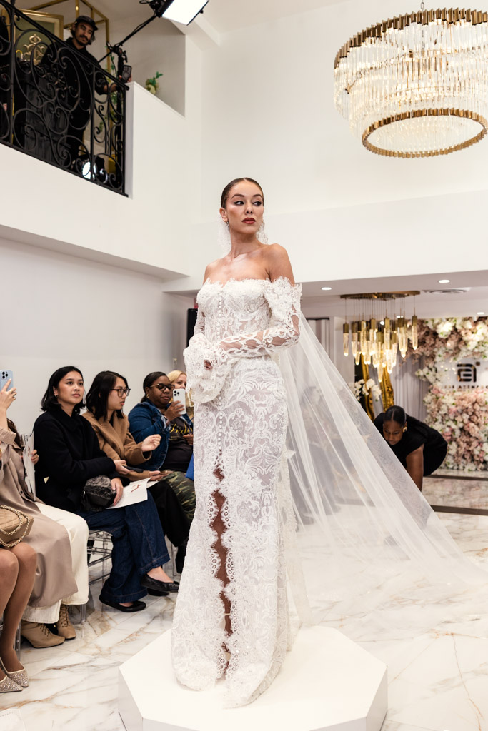 A model in a lacy off-shoulder wedding gown stands on a platform as an audience watches in a bright room at New York Bridal Fashion Week.
