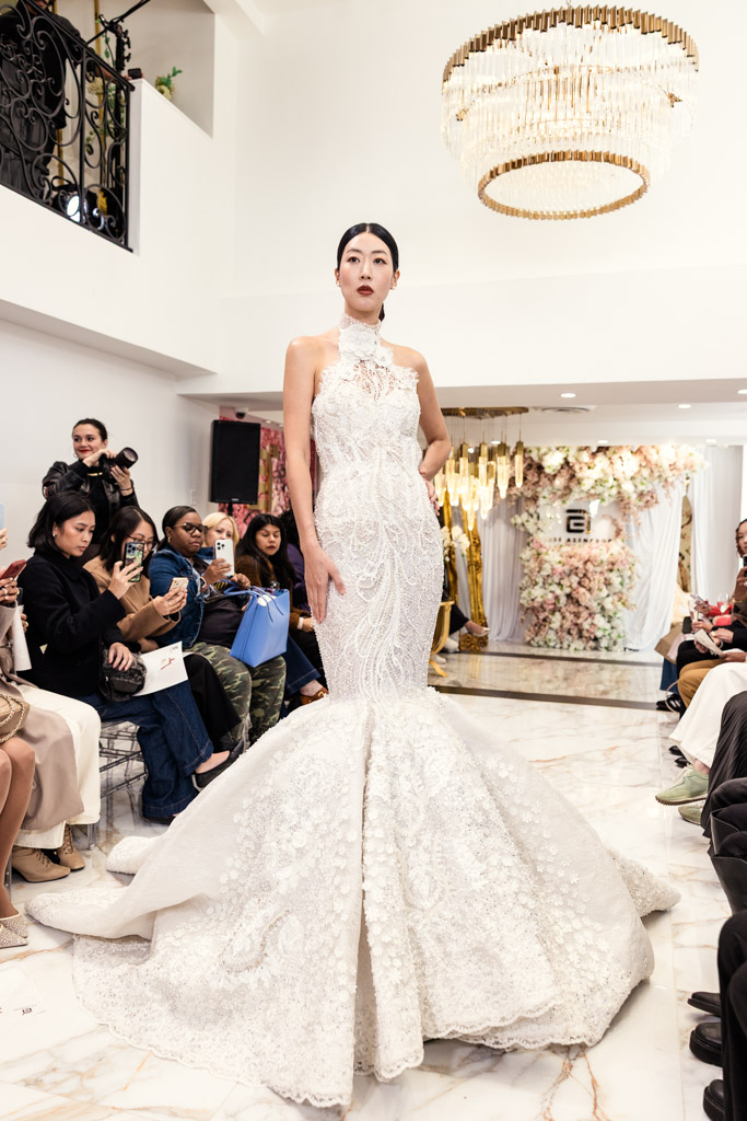 A model in a white mermaid wedding dress poses on the runway at New York Bridal Fashion Week, surrounded by seated spectators.