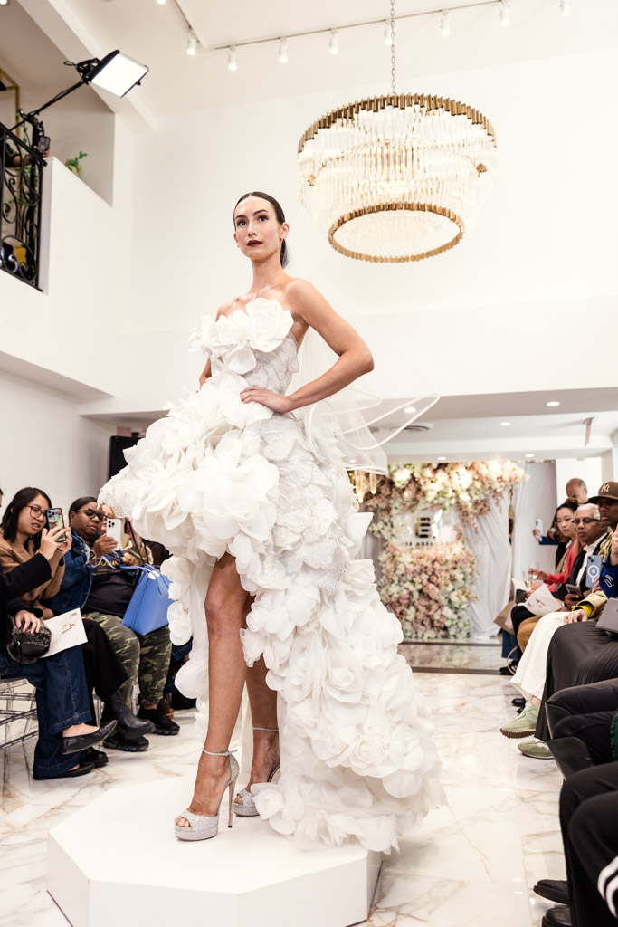 A model in a dramatic white floral gown poses on a runway platform at an indoor fashion show during New York Bridal Fashion Week.