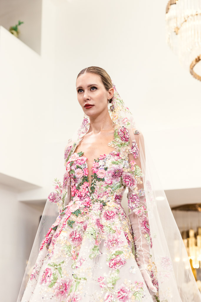 A model in a floral, pastel gown with a matching veil stands under a chandelier in a bright room, capturing the elegance of New York Bridal Fashion Week.