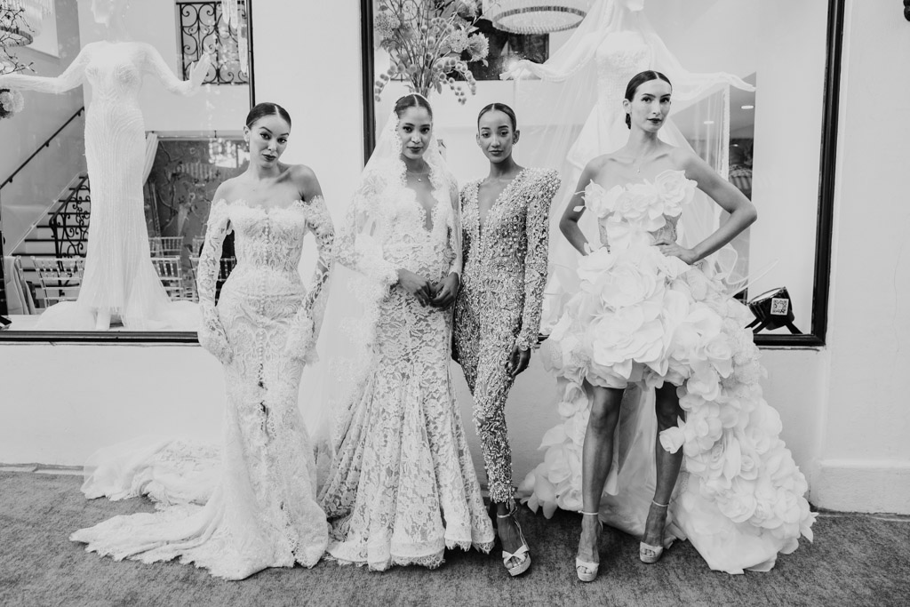 Four women in elegant bridal gowns and jumpsuits pose together outside a bridal shop in black and white, capturing the chic spirit of New York Bridal Fashion Week.