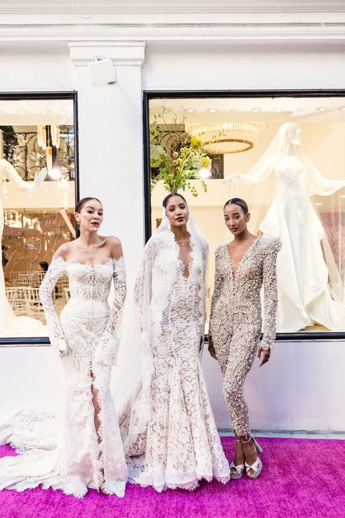 Three women in ornate bridal gowns pose on a pink carpet in front of a bridal shop window display, capturing the elegance of New York Bridal Fashion Week.