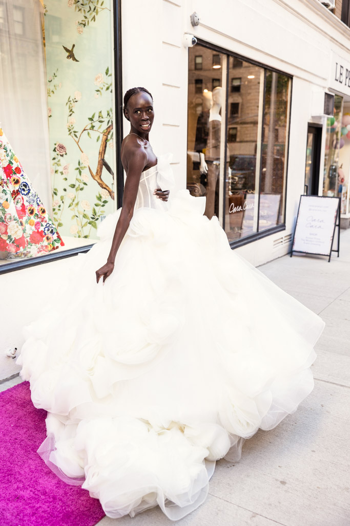 A woman in a voluminous white wedding gown stands on a sidewalk next to a boutique window display, embodying the elegance of New York Bridal Fashion Week.