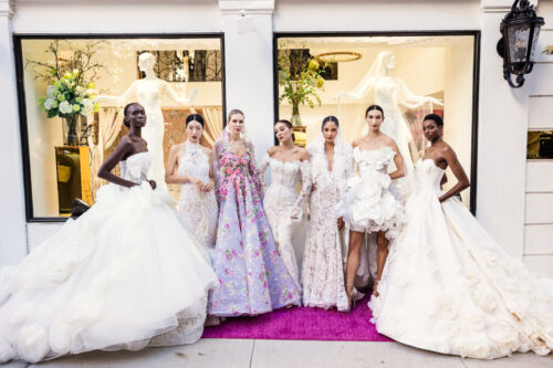 Seven models in elaborate wedding dresses pose on a purple carpet outside a boutique with large display windows, capturing the elegance of New York Bridal Fashion Week.