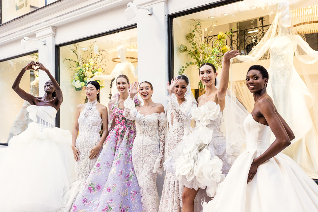 Six women in elegant, ornate wedding dresses pose and smile in front of a bridal shop window display, capturing the glamour of New York Bridal Fashion Week.