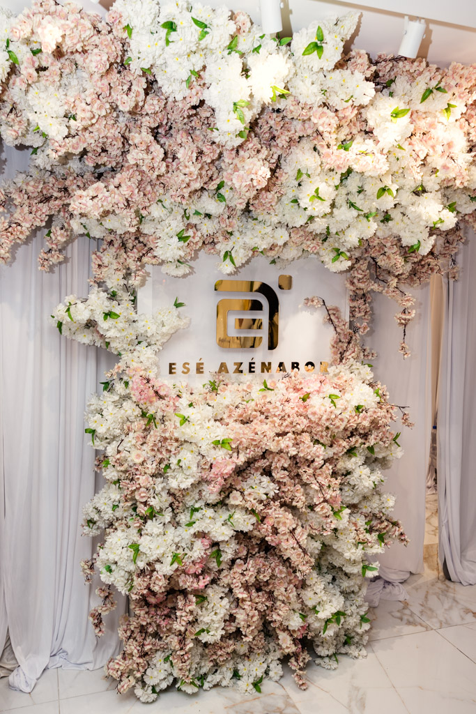 A floral wall with pink and white flowers surrounds a gold Esé Azénabor logo on a white backdrop, creating a stunning display at New York Bridal Fashion Week.