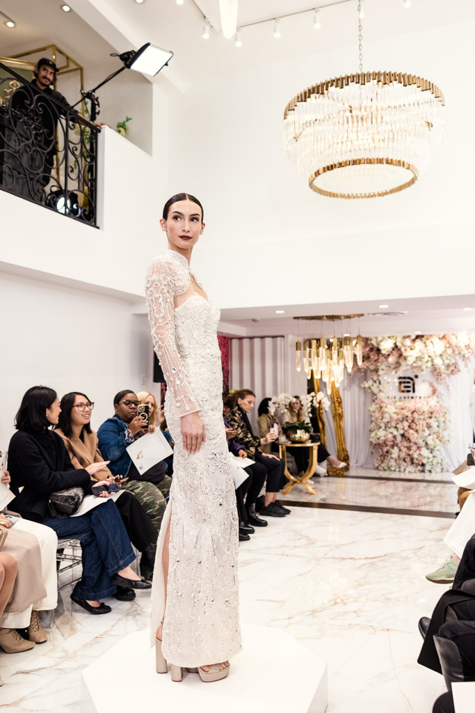 A model in a white lace gown poses on a platform at an indoor New York Bridal Fashion Week show before a seated audience.