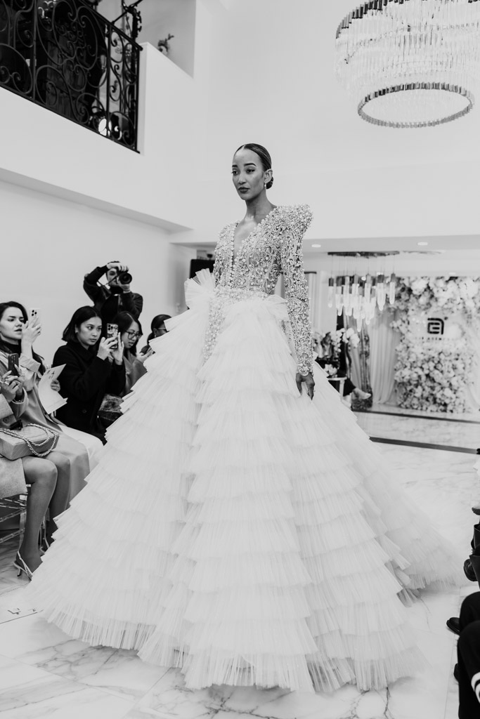 A model in a ruffled, floor-length gown graces the runway at New York Bridal Fashion Week as photographers capture the moment.
