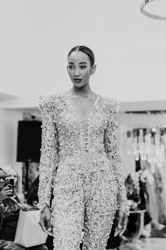 A model walks a runway in an ornate, beaded jumpsuit with intricate detailing at New York Bridal Fashion Week. Black and white photo.