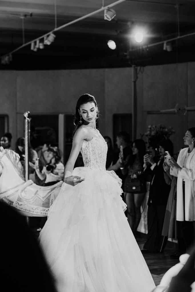 A model in a bridal gown walks on the runway at New York Bridal Fashion Week, while the audience watches and takes photos.