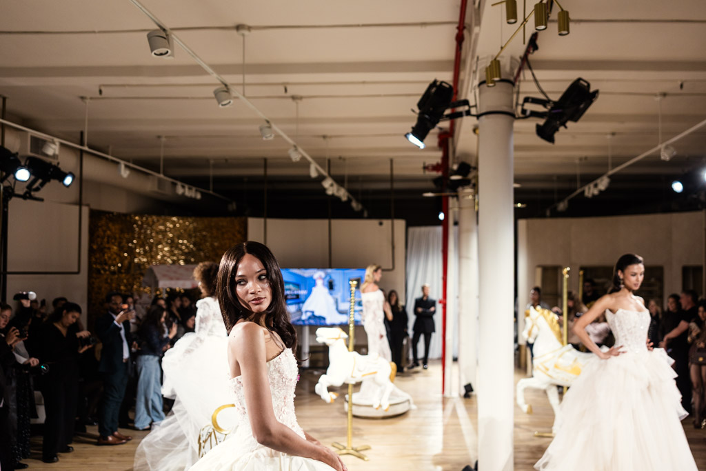 A model in a white gown poses on a runway at New York Bridal Fashion Week, with an audience and carousel horses in the background.