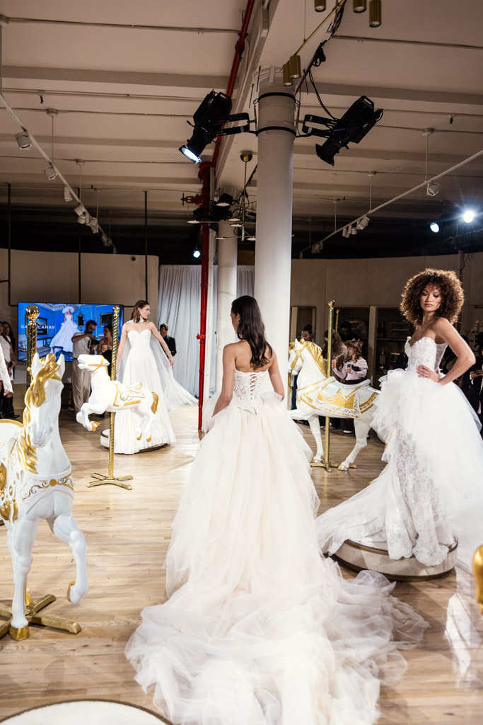 Models in white bridal gowns stand among decorative carousel horses in a brightly lit indoor setting at New York Bridal Fashion Week.